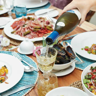 White wine is poured into a crystal glass as guests enjoy a feast, with plates of charcuterie, lentils, salad and shellfish.