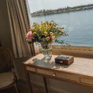 Angled image aboard the Pivoine of a console table with a flower vase and box next to a window overlooking a vast waterway.