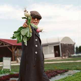 Chef Dominique Crenn, in dark glasses and a flat cap, stands in a vegetable farm, holding up a bunch of fresh-pulled turnips.