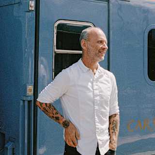 A man with tattoos wearing a white shirt stands and smiles in front of a blue train carriage on a sunny day.
