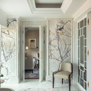 The open double doors of a luxurious and spacious marble bathroom give a glimpse of an elegant sitting room beyond