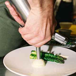 A chef’s hands pipe a creamy purée onto a white plate next to grilled asparagus spears, creating a restaurant-style presentation on a stainless steel surface.