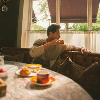 Seated behind a breakfast table with cups and pastries, a man sips coffee as he gazes out of the window onto Pont Street.