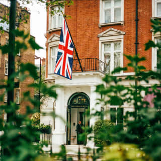 View from Cadogan Place Garden across foliage and over railings to the hotel's white portico entrance, with Union Jack above.