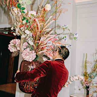 A man in a red velvet jacket carries a huge red glass vase of big blooms and tall stems in shades of pink, seen from behind.