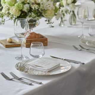 Close-up of an elegant table setting, with porcelain plate, cutlery, wineglasses and large cream and green floral display.