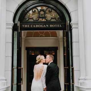 A blonde bride with a backless dress kisses her groom on the marble stoop of the elegant entrance to The Cadogan Hotel