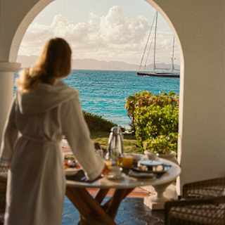 A sailing boat sails by on the azure sea, seen through a patio arch past a robed woman at a breakfast table in soft-focus.