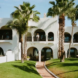 View from the garden to a white, Moroccan-style, two-level villa with arched colonnade balcony and terrace, flanked by palms.