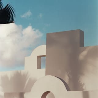 A detail of a bright white villa in half shade looks like geometric art with angles and shapes stamped on a blue, cloudy sky.