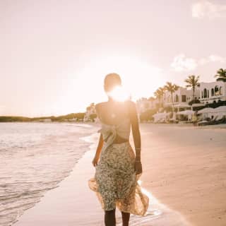 Dame in einem Sommerkleid, die bei Sonnenuntergang barfuß den Strand entlang geht