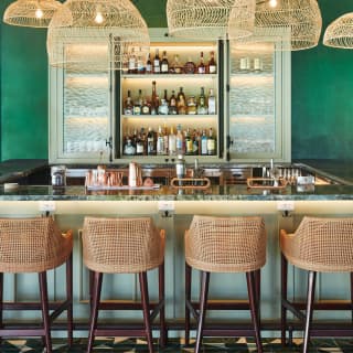 Rattan backed barstools line a highly polished bar. On the rear wall, open glass doors reveal well-stocked shelves of liquor