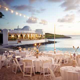 White circular tables on a beach at sunset, lit with fairy light