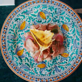 A colourful, ornate plate with a slice of yellow omelette on top of a pile of thinly sliced ham, set on a dark green table.