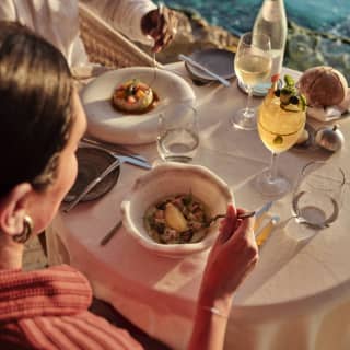 A couple share a meal with white wine and a cocktail at a Pimms terrace table overlooking the ocean, seen from a high angle.