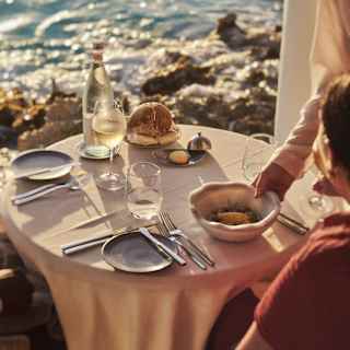 A waiter places a bowl of Fresh Local Catch in front of a diner who enjoys a sunny waterfront meal with white wine at Pimms.