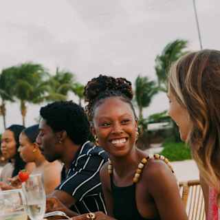 A woman turns towards the camera to smile at a friend in a view of guests enjoying a private beach dinner, seen in close-up.
