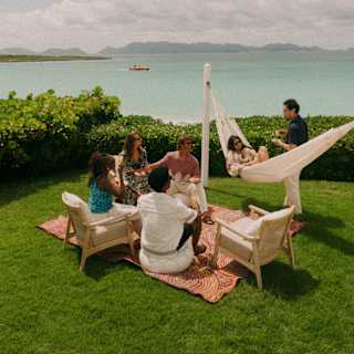 Guests chat in a lush green sea-view garden, with two on chairs, two on a blanket, one in a hammock and another standing.