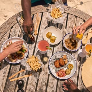 Diners' hands reach for grilled crayfish, corn, fries, and tasty plates at a Cap Shack table in the sand, seen from above.