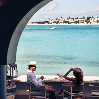 A smiling couple sitting at a restaurant counter table on an open-air terrace overlooking the Caribbean sea