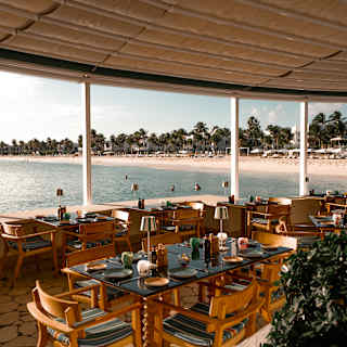 Open-air restaurant with wooden tables and chairs overlooking a sandy beach and calm sea. Palm trees and sun loungers are visible in the background along the shoreline under a bright, partly cloudy sky.