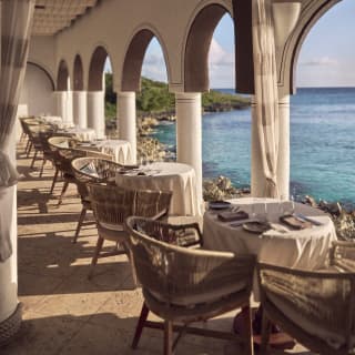 View through the seafront colonnade terrace at Pimms restaurant, where a row of six round tables for two are laid for lunch.