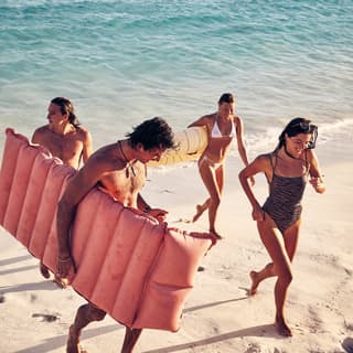 Four smiling guests in swimsuits jogging from the sea towards the beach holding lilos and floats