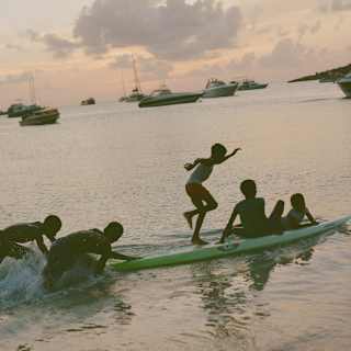 In the sunset light, five local children play with a green surfboard in the shoreline waters, with yachts moored behind.