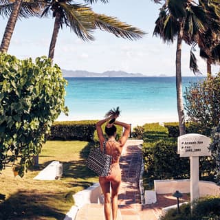 A lady pulling her hair into a ponytail as she strolls down a villa path towards a beach