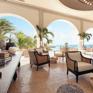 Greco Moorish arches frame picture perfect white beaches, palm trees and blue seas beyond a lounge of dark wood armchairs