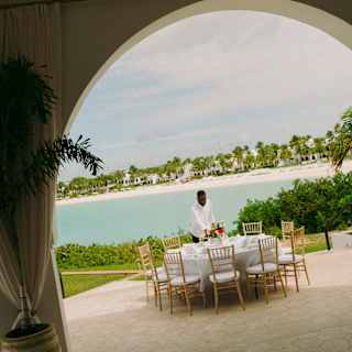 Angled image of a male staff member at a round table set for ten on a sea-view terrace, seen from inside the event pavilion.
