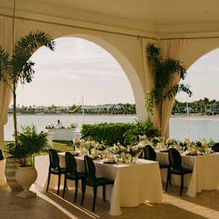 Two tables with white cloths and floral centrepieces await wedding guests in the event pavilion with stunning bay views.