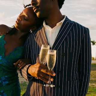A woman in a blue dress rests her head on the shoulder of a male friend in a pinstripe suit holding two glasses of Champagne.