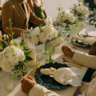 Suited guests sits at a wedding table with white flower displays, dark plates, tealights and green glassware, in close-up.