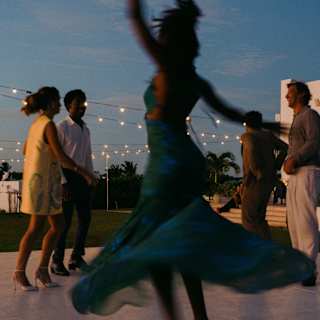 A woman dances in soft-focus foreground, dress fanning around her, on a dancefloor with light strings below a darkening sky.