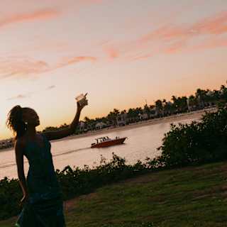 A woman holding up a drink is silhouetted against a pink sunset sky that tints the bay behind, where a small boat cruises by.