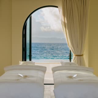Two beds with crisp linen face the wide blue view through an open arched window in the Imperial VIP double treatment room.