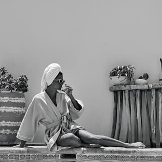 Black and white photo of a lady in a bathrobe posing on a poolside and sipping from a glass