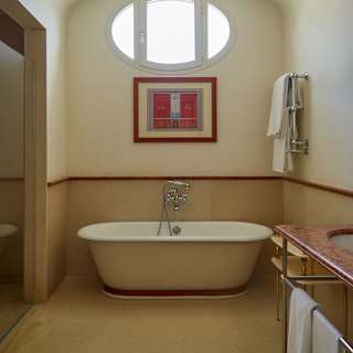 An oval window in an alcove, above a painting and freestanding bathtub, glows with light in the elegant bathroom.