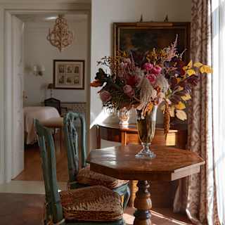 A hexagonal wood table with flowers catches light from the window in a view from the living room through to the bedroom.