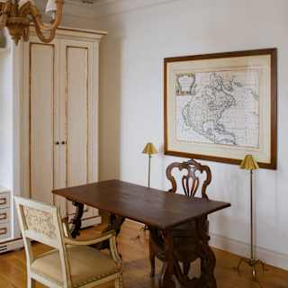 A wooden table with two antique chairs, one cream, one natural, seen in a room with a framed map in Villa Margherita.