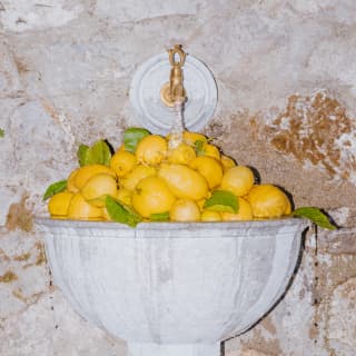 A mountain of lemons fills a marble sink set into a stone exterior wall as a running  tap overflows