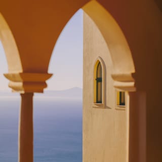 A pointed Moorish arch in Naples yellow is turned honey gold by the low sun as it reveals ocean views of deepest azure