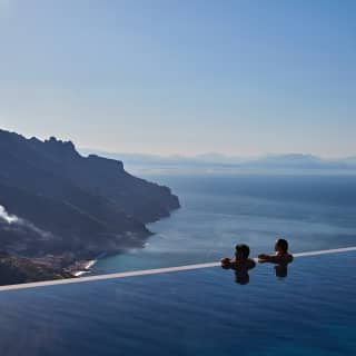 A man and a woman rest at the edge of an infinity pool, gazing at the view that plunges down steep cliffs below them