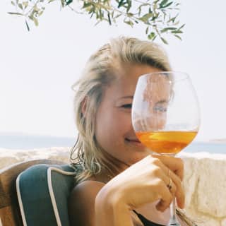 A young woman looks at the camera through the top of a glass of Aperol as she sits in the shade of an olive tree