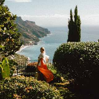 A blonde woman in a white top and breeze-blown orange skirt stands at a viewpoint in gardens overlooking the dramatic coast.