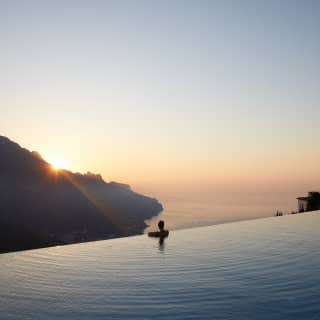 The last cathedral rays of sunshine dip below the Lattari Mountains as a woman gazes on from an infinity pool’s edge