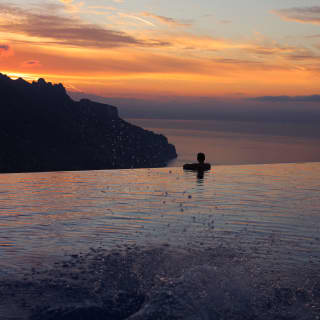 A swimmer rests at the edge of infinity pool watching the silhouetted coastline melt into the purple haze beneath a sunset.