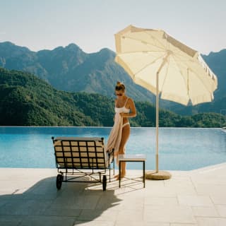 A woman in a white bikini stands with a towel next to her poolside lounger, beneath a parasol, with a mountainous backdrop.