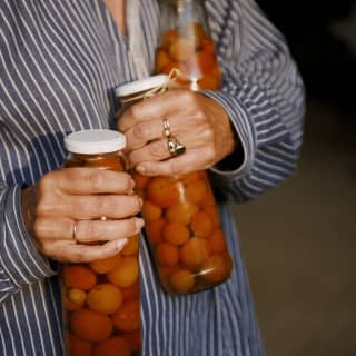 A woman in a striped shirt carries three tall jars of pickled cherry tomatoes, one in each hand and one under her left arm